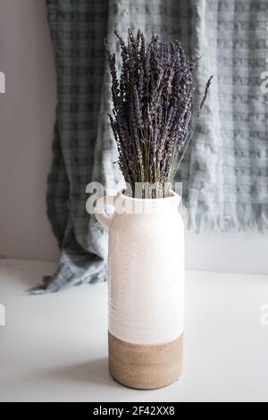 Bouquet von trockenen Lavendelblüten in einer rustikalen Tonvase über einem weißen Tisch mit einer aufgehängten, verschwommenen, türkisfarbenen Decke im Hintergrund. Stockfoto