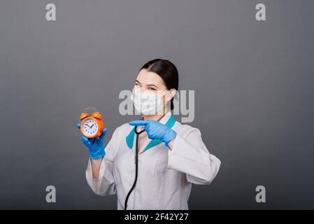 Lächelnde Ärztin Frau in Maske zeigt auf Uhr, Zeit zu impfen, medizinische Konzept Stockfoto