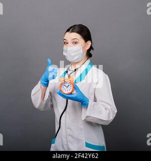 Lächelnde Ärztin Frau in Maske zeigt auf Uhr, Zeit zu impfen, medizinische Konzept Stockfoto