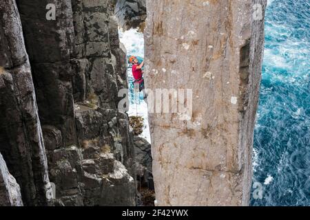 Abenteuerlustige Menschen klettern eine freiliegende Felssäule mit Meeresklippen und das Meer im Hintergrund in der Totem Pole, Cape Hauy, Tasman National Park, Tasmanien, Australien. Stockfoto