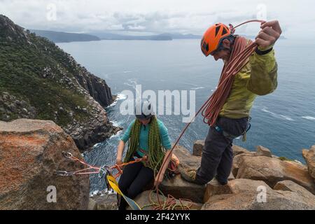 Abenteuerlustige Paare lachen beim Seilrollen, während sie an einem bewölkten Tag in Tasmanien, Australien, die freiliegenden Meeresklippen von Cape Raoul erklimmen. Stockfoto