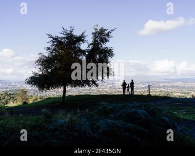 Eine dreiköpfige Familie, die neben einem Baum steht und die Aussicht genießt und vom Naturpark Cathkin Braes aus die Stadt Glasgow überblickt. Stockfoto