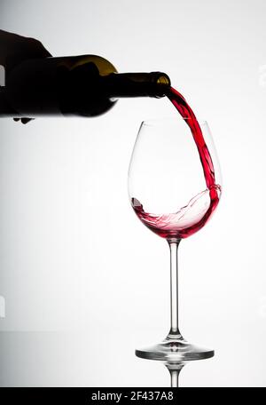 Vertikale hinterleuchtete Abbildung einer Flasche, die ein Glas füllt Rotwein fängt den Moment ein, in dem der Wein gerade gekommen ist In Kontakt mit dem Glas Stockfoto