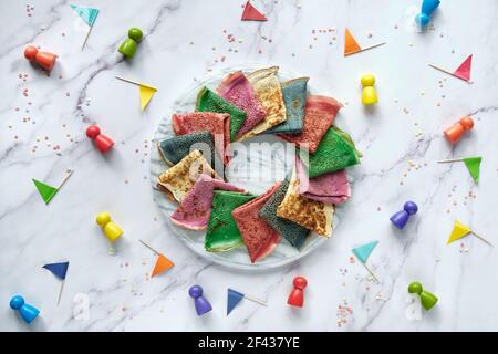 Rainbow Party, Tischaufbau. Lebendige Crepes auf Marmortisch mit Holzfiguren und Papierfahnen. Kreatives Essen. Regenbogen als Symbol des Mitgefühls Stockfoto