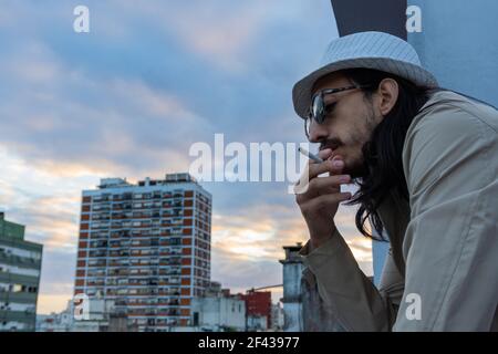 Junge Hipster mit langen Haaren Rauchen Zigarette. Mann mit Hut und Sonnenbrille Rauchen Tabak auf dem Balkon mit Blick auf die Stadt beobachten den Sonnenuntergang. l Stockfoto