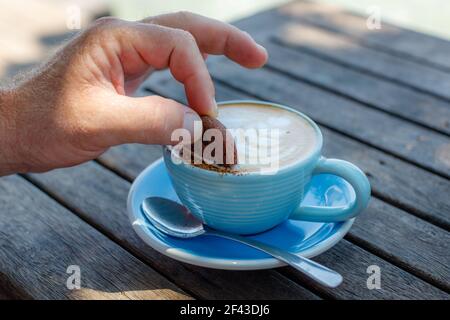 Männliche Hand taucht einen Keks in eine blaue Tasse Cappuccino und steht auf einem Holztisch in einem Café. Mit Platz. Stockfoto