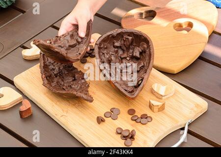 Kind nimmt die Hälfte der dunklen Schokolade Osterei mit knackigen Mandeln. Umgeben von Schokoladentafeln und Holzherzen. Stockfoto