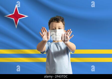 Kleiner weißer Junge in einer Schutzmaske auf dem Hintergrund der Flagge von Aruba. Macht ein Stoppschild mit seinen Händen, zu Hause bleiben Aruba. Stockfoto