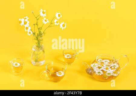 Drei Tassen Kamillentee, transparente Teekanne und Vase mit Gänseblümchen-ähnlichen Blumen auf gelbem Hintergrund. Kamille Tee Vorteile Ihr Gesundheitskonzept. Stockfoto