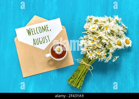 Willkommen August Text auf weißer Karte in Handwerk Umschlag, Blumenstrauß weißen Kamillen Blumen und Tasse Tee auf blauem Hintergrund. Konzept Hallo august. Greetin Stockfoto