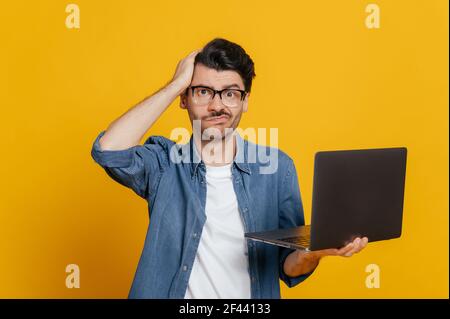 Frustriert unrasiert kaukasischen Kerl in Brille und in Denim-Shirt, hält einen Laptop, schaut verwirrt auf die Kamera, hält den Kopf, ohne Stimmung, steht auf einem isolierten orangen Hintergrund Stockfoto
