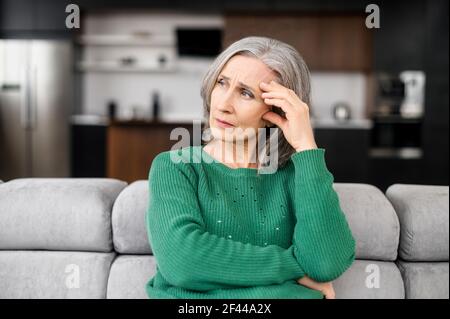 Verärgert depressive ältere grauhaarige Dame trägt grünen Pullover berühren Tempel, sitzen auf dem Sofa, Blick in das Fenster, Gefühl besorgt und gestresst über die Zukunft und denken über eigene Probleme Stockfoto