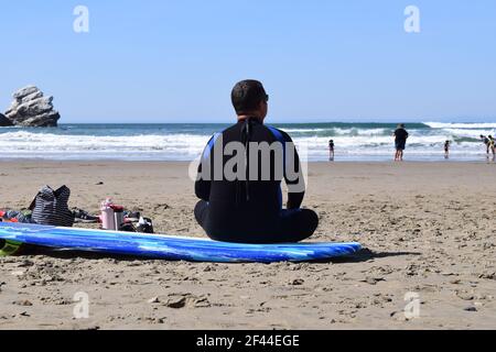 FRESNO, USA - 28. Feb 2021: Ein Foto eines männlichen Surfers, der auf seinem blauen Surfbrett am Strand sitzt und in den Ozean blickt Stockfoto