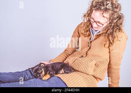 Detail einer schönen Frau mit Brille und lockig braunem Haar, lächelnd sitzend, mit einem fast schlafenden Jack Russel Terrier Welpen auf den Beinen. Zoll Stockfoto