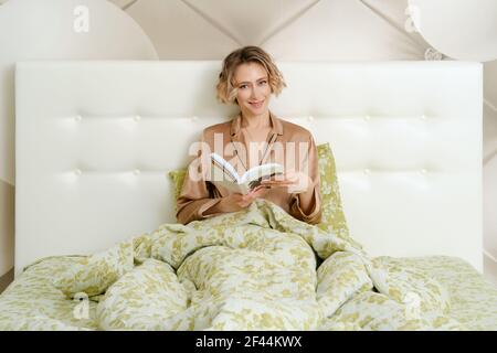 Clevere junge Frau sitzt im Bett unter der Decke mit Ein Buch in den Händen Stockfoto