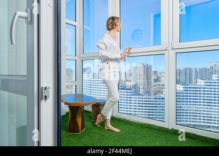 Barfußfrau steht auf dem Balkon ihrer neuen Wohnung Und bewundert die Aussicht auf die Stadt aus dem Fenster Stockfoto
