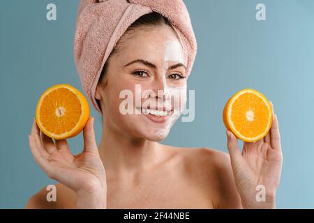 Schöne junge Frau trägt Badetuch auf dem Kopf zeigt Orangen, trägt kosmetische Gesichtsmaske Stockfoto