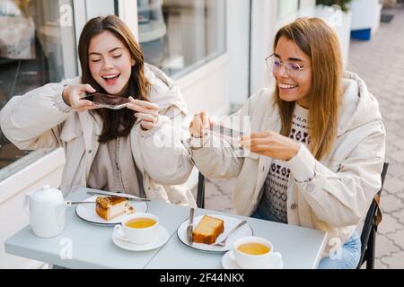 Junge fröhliche Freundinnen sitzen im Café im Freien und nehmen Bilder vom Essen mit Handy Stockfoto