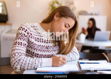 Lächelnd charmante Student Mädchen Hausaufgaben machen, während im Café sitzen In Innenräumen Stockfoto