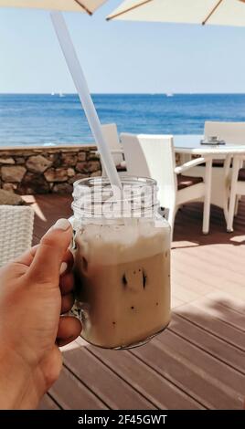 Eine vertikale Aufnahme einer weiblichen Hand, die ein Eis hält Kaffee mit Meereshintergrund Stockfoto