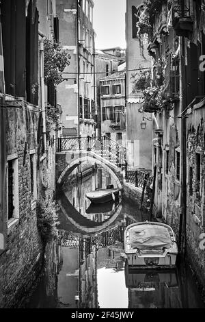 Seitenkanal in Venedig mit kleiner Brücke und festverankerten Booten, Italien. Schwarz-Weiß-Stadtfotografie, venezianisches Stadtbild Stockfoto