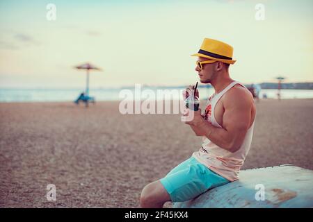 Der junge, gutaussehende sportliche Mann in heller Kleidung, Strohhut und Sonnenbrille sitzt am Meer, trinkt einen erfrischenden Cocktail und genießt seinen Urlaub. Stockfoto