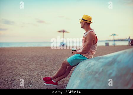 Der junge, gutaussehende sportliche Mann in heller Kleidung, Strohhut und Sonnenbrille sitzt am Meer, trinkt einen erfrischenden Cocktail und genießt seinen Urlaub. Stockfoto