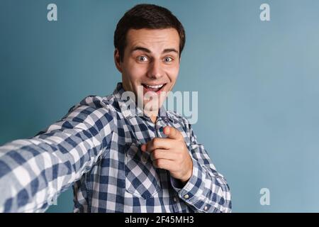 Attraktiver lächelnder junger Mann im karierten Hemd, der Selfie auf blauem Hintergrund macht und mit dem Finger auf die Kamera zeigt Stockfoto