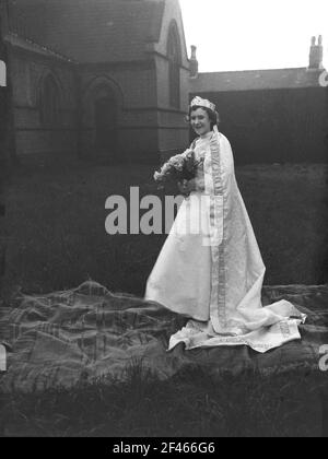 1956, historisch, ein älteres Teenager-Mädchen, das draußen auf dem Gelände einer Kirche steht, in einem langen Kleid und einem aufwendigen Spitzenkleid mit Pfad und einem Blumenstrauß, bevor es an der Parade zum 1. Mai teilnimmt, Ein beliebter Tag der Frühlingsfeier in vielen Teilen Nordenglands in der Ära der Tihs. Auf ihrem Kopf eine Krone, die zur diesjährigen May Queen gewählt wurde. Der erste Mai, allgemein bekannt als Maifeiertag, ist ein gefeierter Frühlingstag, der den Beginn des Sommers markiert und auf das uralte Fest der römischen Blumengöttin Flora zurückgeht. Stockfoto