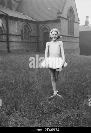 1956, historisch, ein junges Mädchen, das draußen auf dem Gras auf dem Gelände einer Kirche in einem kurzen Ballettkleid steht, bevor es an der Parade zum 1. Mai teilnimmt, einem beliebten Feiertag, der den Beginn des Sommers markiert. May First, allgemein bekannt als May Day, ist eine alte Tradition, die auf die Feier der römischen Blumengöttin Flora zurückgeht. Stockfoto