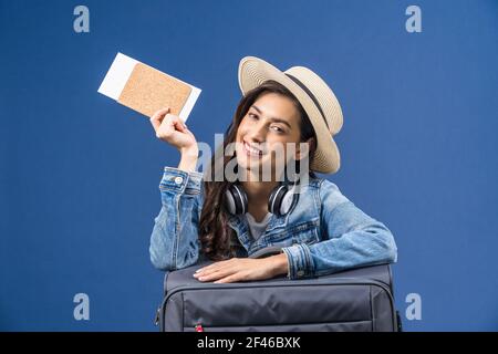 Glückliche junge asiatische Frau mit Pass mit Bordkarte und präsentiert auf blauem Hintergrund, Kopfhörer und Gepäck, Urlaub und Reisekonzept Stockfoto