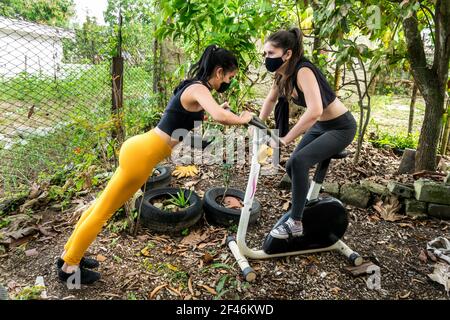 Zwei junge kubanische Frauen arbeiten zusammen draußen in einem verlassenen Hinterhof, sie sind mit einem statischen Fahrrad Stockfoto