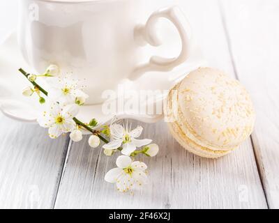 Nahaufnahme von beigefarbenen Makronen und Frühlingskirschblüten gegen weiße elegante Teetasse auf einem weißen Holztisch. Süßes Frühstück mit köstlichem almon Stockfoto