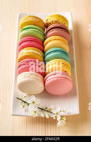 Set aus bunten Makronen und Frühlingskirschblüten auf einer weißen rechteckigen Platte gegen Natupal Holztisch. Leckere französische Mandelmeringue. Stockfoto
