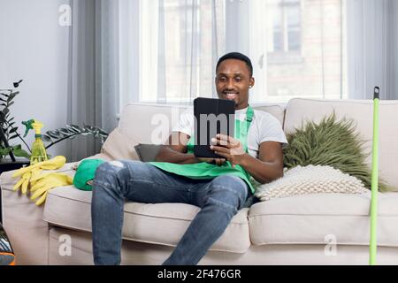 Lächelnder afro-amerikanischer Hausmeister sitzt auf der Couch mit Tablet Stockfoto