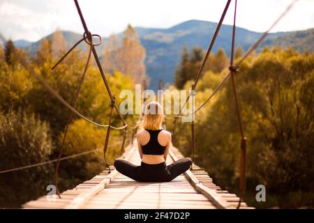 Rückansicht eines jungen ruhigen Mädchens, das Yoga-Meditation praktiziert, in Lotushaltung auf der Brücke über den Fluss sitzt. Wohlfühlkonzept. Stockfoto