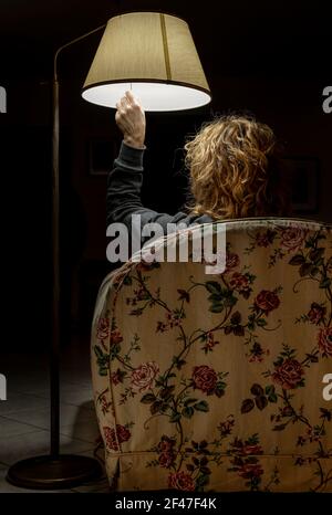 Eine blonde Frau, die von hinten geschossen wird, sitzt in einem Sessel und beleuchtet eine antike Lampe durch Ziehen einer Schnur Stockfoto