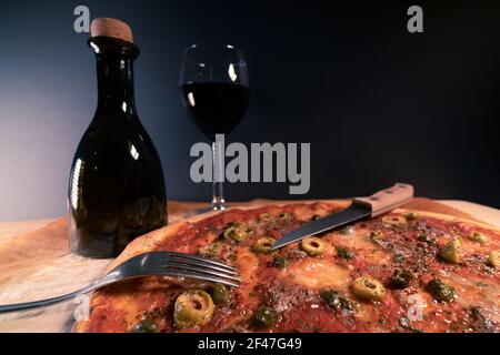 Frisch gebackene, hausgemachte Pizza Napoli auf Backpapier direkt nach dem Ausgehen aus dem Ofen gelegt. Ein Glas Rotwein und eine Flasche Olivenöl. Oliven, Sardellen Stockfoto