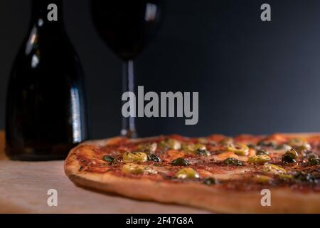 Frisch gebackene, hausgemachte Pizza Napoli auf Backpapier direkt nach dem Ausgehen aus dem Ofen gelegt. Ein Glas Rotwein und eine Flasche Olivenöl. Oliven, Sardellen Stockfoto