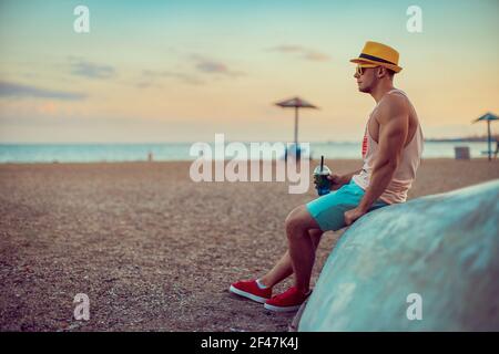 Der junge, gutaussehende sportliche Mann in heller Kleidung, Strohhut und Sonnenbrille sitzt am Meer, trinkt einen erfrischenden Cocktail und genießt seinen Urlaub. Stockfoto