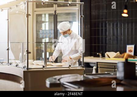 Junger Koch in Gesichtsmaske, der an der Präsentation des Essens arbeitet Stockfoto