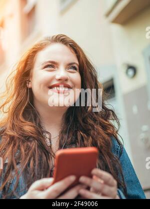 Lächelnd rot lockig lange Haare kaukasischen Teenager-Mädchen zu Fuß auf der Straße und Surfen im Internet mit dem modernen Smartphone. Moderne Leute mit Techno Stockfoto