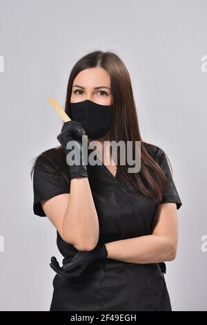 Frau Kosmetikerin in schwarzem Anzug und Gesichtsmaske hält Enthaarung Stick Stockfoto