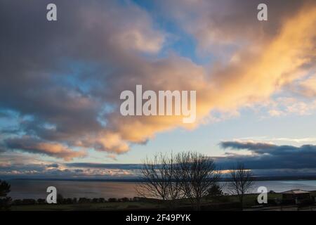 Silhouettierte Winterbäume auf einer Esplanade vor einer Bucht von Wasser, während der Sonnenuntergang die Wolken erhellt. Stockfoto