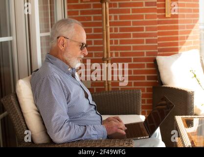 Alter Mann mit Bart glücklich auf der Terrasse arbeitet von Zu Hause mit einem Laptop Stockfoto