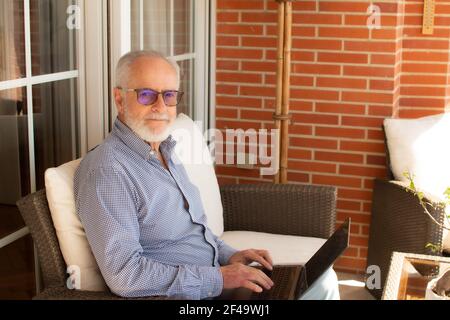 Alter Mann mit Bart glücklich auf der Terrasse arbeitet von Zu Hause mit einem Laptop Stockfoto