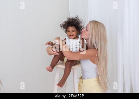 Porträt der jungen blonden kaukasischen Mutter und afroamerikanischen glücklich lockig Tochter auf weißem Hintergrund. Ein Mädchen steht auf einem Holzstuhl, eine Frau umarmt, Stockfoto