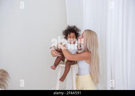 Porträt der jungen blonden kaukasischen Mutter und afroamerikanischen glücklich lockig Tochter auf weißem Hintergrund. Ein Mädchen steht auf einem Holzstuhl, eine Frau umarmt, Stockfoto