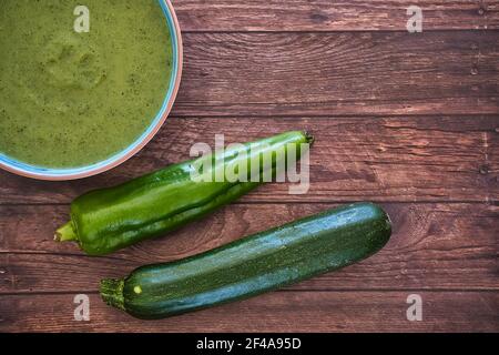 Frische Gemüsesuppe aus Erbsen auf einem Teller auf braunem Holztisch mit Gurken und grünem Pfeffer, Draufsicht mit Platz für Text. Lecker Stockfoto