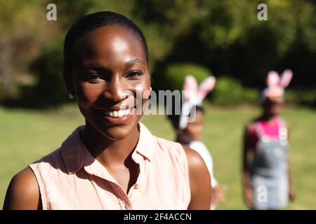 Porträt einer afroamerikanischen Mutter, die lächelt und Kinder trägt, die Hase tragen Ohren im Hintergrund Stockfoto